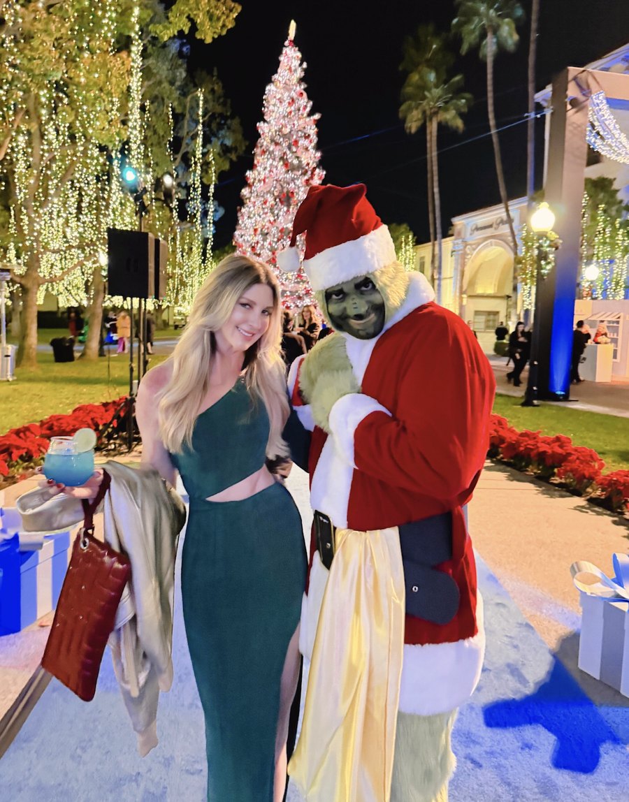 Kelsy Elle in a green gown at a holiday party, beside a costumed Grinch, under lit Christmas trees